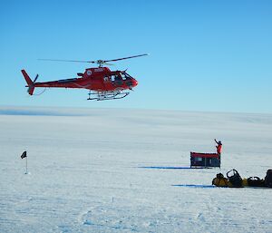 A helicopter is approaching a cage pallet of equipment on the glacier. A person is there ready to receive the sling line.