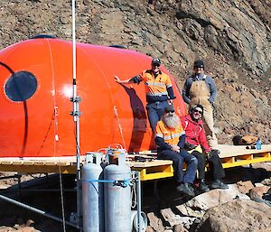 A group shot of Vas, Val, Ed and Rhys leaning against the newly erected melon hut. It looks bright, shiny and very very orange.