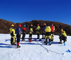 The lads are standing on the ice, learning how to anchor in rope lines in order to abseil down the ice cliff.