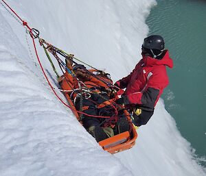 Sharky is strapped into the stretcher, which is being pulled up the ice cliff using ropes and pulleys. A rescuer is with him.