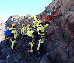 The SAR team are standing below a rock cliff while the FTO shows them how to install anchor points into the rocks.