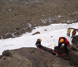Two people are on the rockface with the patient in the stretcher as it is being hauled up to safety.