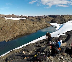 Sharky, Gideon and Darren are sitting on a high rocky outcrop, overlooking a long lake with snow drift around it.