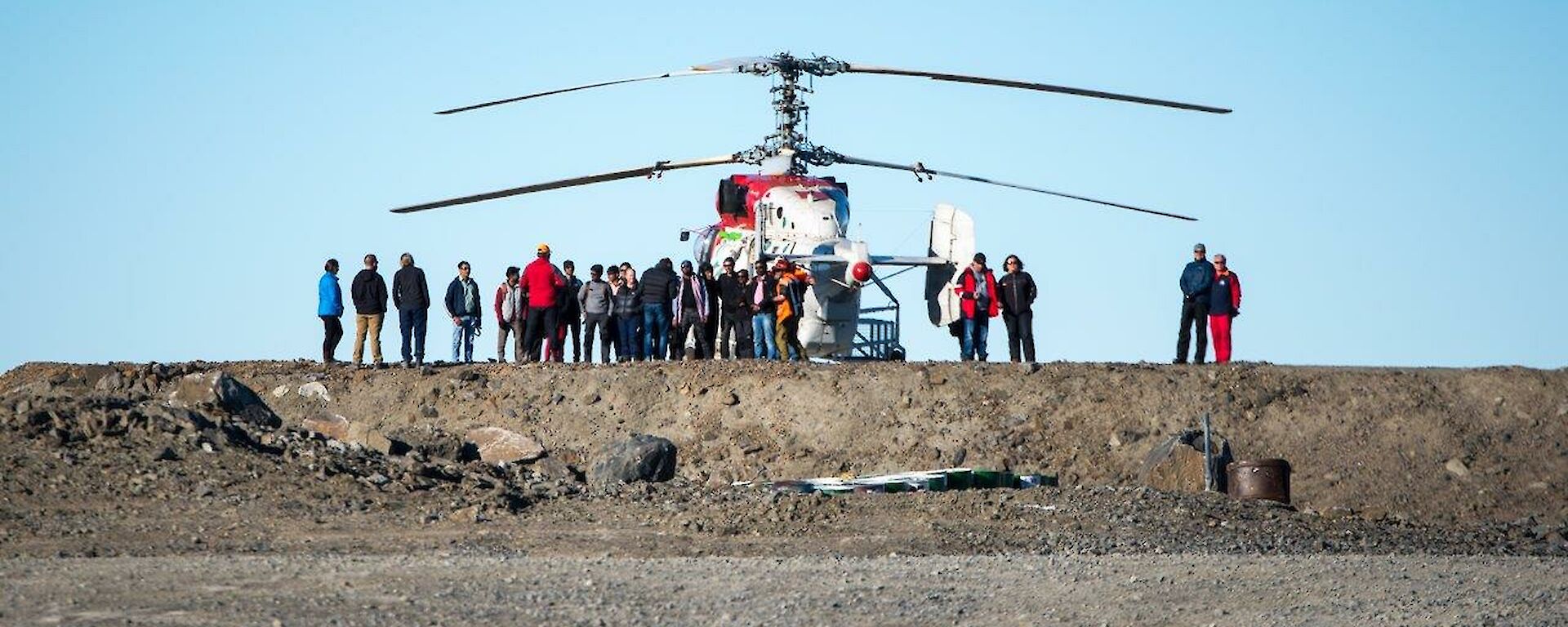 Many people are seen in silhouette against the blue sky and Kamov helicopter, sitting on the helipad.