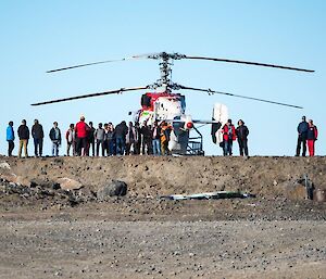 Many people are seen in silhouette against the blue sky and Kamov helicopter, sitting on the helipad.