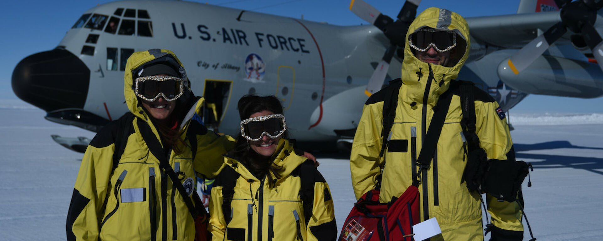 three people dressed in their yellow outer shells with ski googles. They are standing on the Wilkin’s skiway with the Hercules in the background.