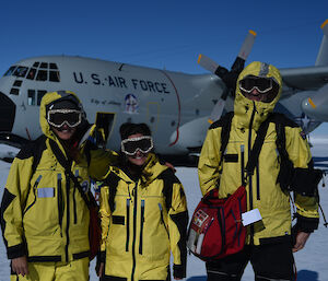 three people dressed in their yellow outer shells with ski googles. They are standing on the Wilkin’s skiway with the Hercules in the background.