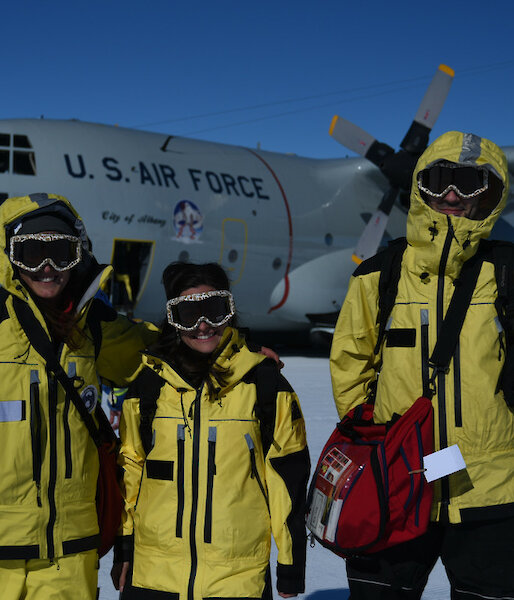 three people dressed in their yellow outer shells with ski googles. They are standing on the Wilkin’s skiway with the Hercules in the background.