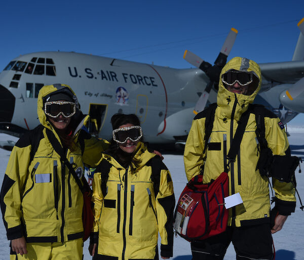 three people dressed in their yellow outer shells with ski googles. They are standing on the Wilkin’s skiway with the Hercules in the background.