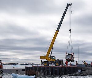 The drill rig is mid air, being lifted by a large crane and about to be put onto a barge and transported to the ship.