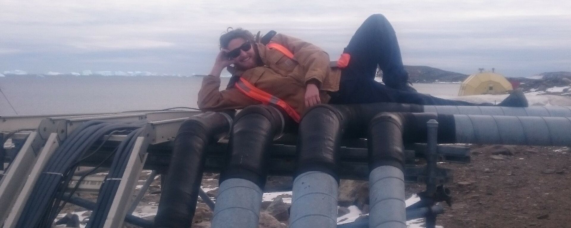 Bryce is lying down on the pipes of the site services. In the background is Prydz Bay and icebergs.