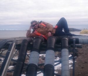 Bryce is lying down on the pipes of the site services. In the background is Prydz Bay and icebergs.
