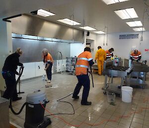 Three people are seen cleaning the floor in the gutted kitchen. Tony is power washing the tiles, Rhys is scubbing with a broom and Kirsten is vacuuming up the water.