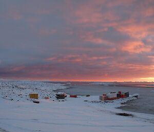 The wharf has a number of shipping containers on it, has a nice cover of snow and there is a pink sunset in the distance.