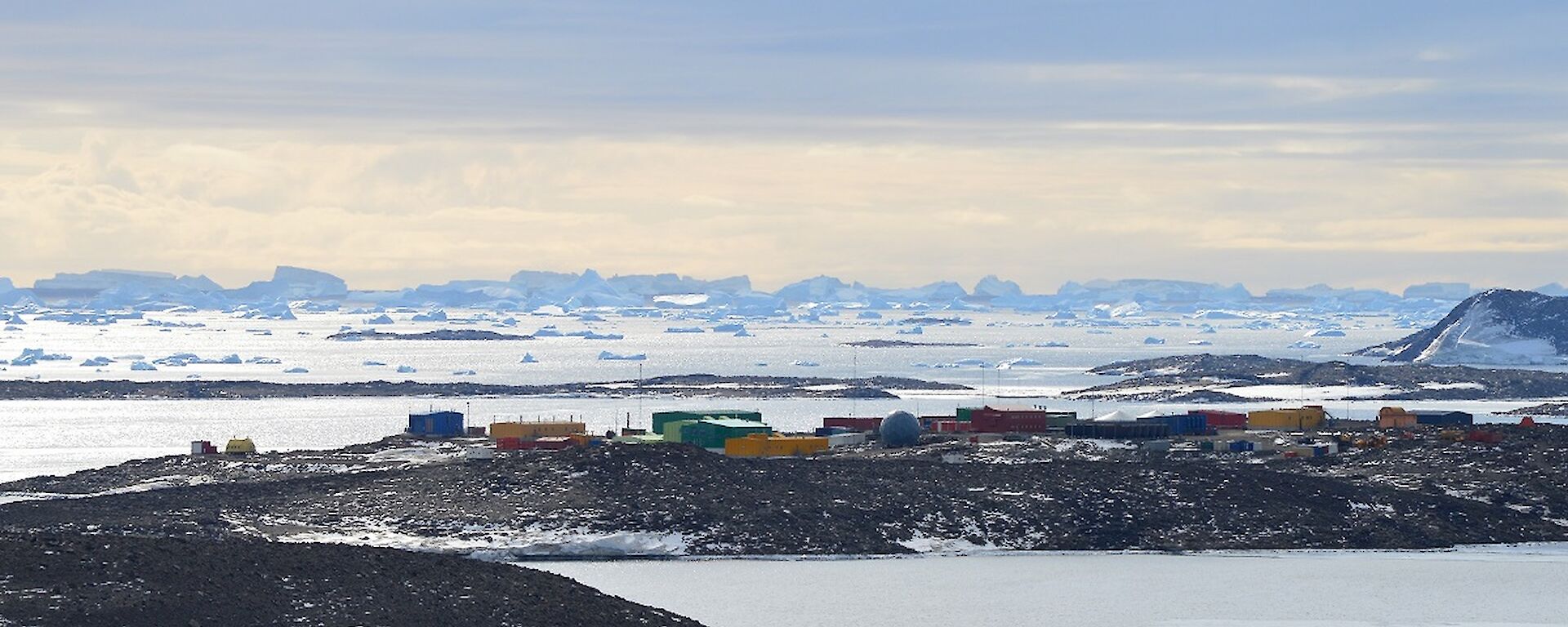 A view from the top of The Lookout. There are brown hills amongst the ice, with colourful buildings in the distance. Behind the station are icebergs on the horizon.