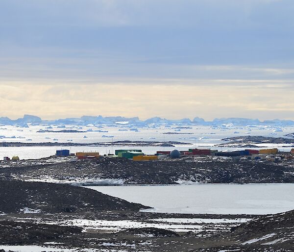 A view from the top of The Lookout. There are brown hills amongst the ice, with colourful buildings in the distance. Behind the station are icebergs on the horizon.