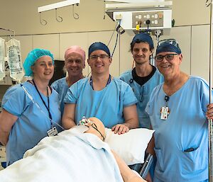 The Lay Surgical Assistants team standing around dressed in blue scrubs, surrounding a dummy patient in Hobart Hospital during training.