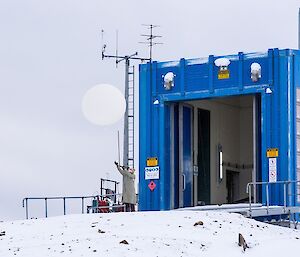 Daleen is dressed in safety gear, holding a weather balloon in front of the blue Met building.