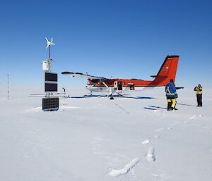 The twin otter plane is parked on the ice shelf. Close by some scientific equipment is sticking out above the ice. This looks like solar panels and a wind turbine. Expeditioners are nearby taking photos.