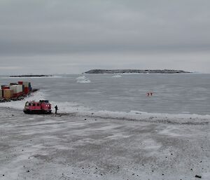 Two expeditioners in bright orange dry suits out on the sea ice. The pink Hägg and an expeditioner are on shore as the support system.