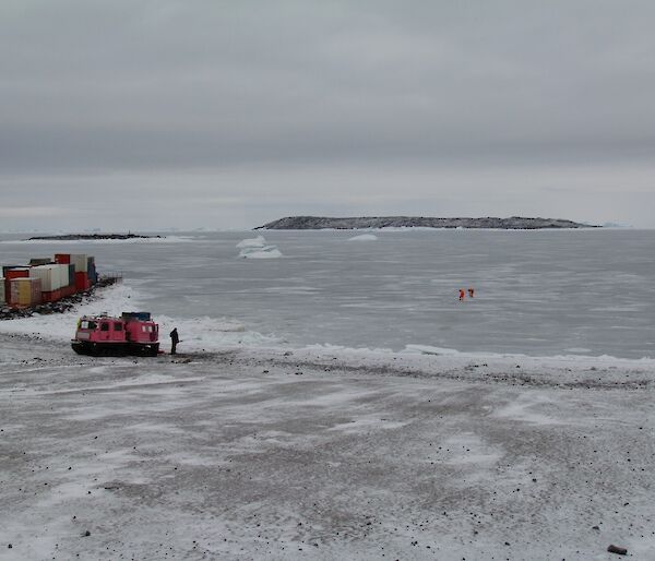 Two expeditioners in bright orange dry suits out on the sea ice. The pink Hägg and an expeditioner are on shore as the support system.