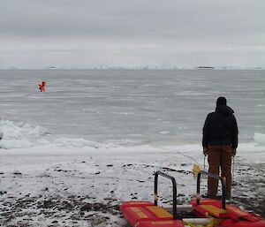 An expeditioner stands on shore watching the boys out on the ice. He holds the belay rope to the boys and has a rescue platform beside him.