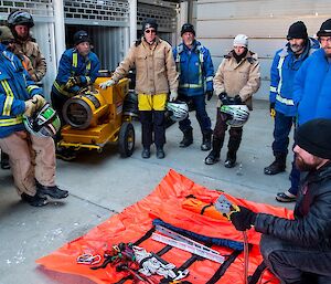 Marc is bend down by an orange tarp, covered in tools. He is showing the team surrounding him, the different equipment used for quad recovery.