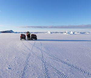 Two quads are parked on the sea-ice. Two people stand between the bikes talking to each other.