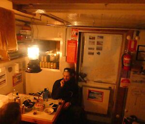Lötter relaxing inside Platcha Hut. He’s sitting at a small table. Maps line the hut wall and a tilly lamp lights the hut.