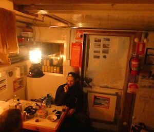 Lötter relaxing inside Platcha Hut. He’s sitting at a small table. Maps line the hut wall and a tilly lamp lights the hut.