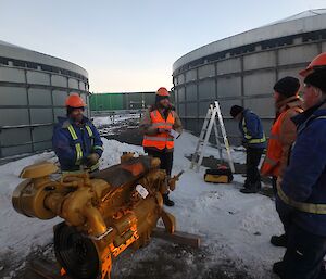 Jock is holding a safety meeting before they start the job. In front of him is the new engine to go into the Powerhouse. Five others are seen in attendance, all with safety gear on.