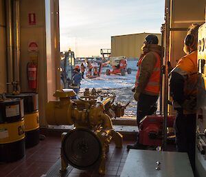 Jock and Barry B2 are standing inside the Powerhouse, looking out to the activities outside. They are waiting for the side panel to be delivered. The new engine is sitting on the Powerhouse floor.