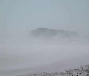 The green utilities building is seen in silhouette behind the thick snow blowing down the road.