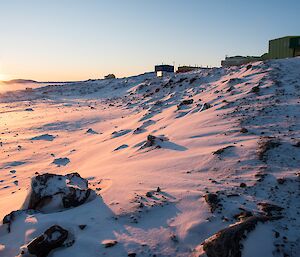 The sun is rising, lighting up the snow to a gentle pink colour out the front of Davis station.