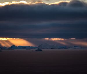 Dramatic sunset over the icebergs as an orange light beams through the dark grey clouds.