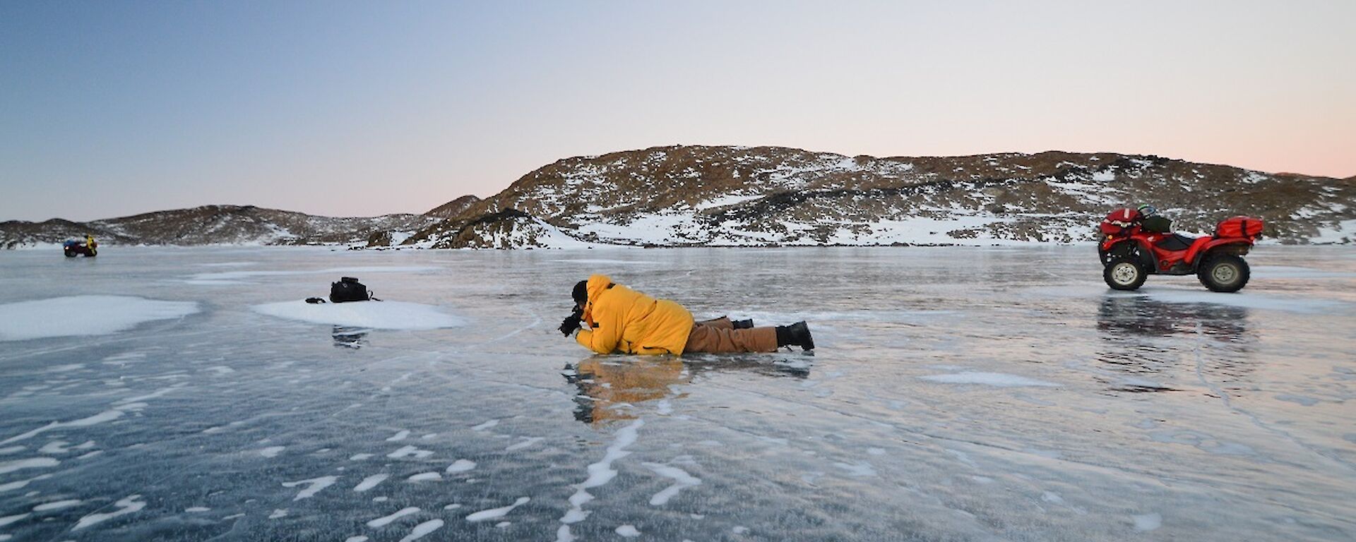 Barry (B1) is lying on the lake ice taking photos of the cracks in the ice. You can see a couple of quad bikes parked on the ice as well. Behind B1 are the Vestfold Hills and twilight skies.