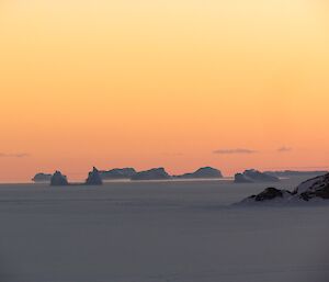 A mandarin orange sky behind the bergs during civil twilight.