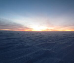A landscape shot of the Antarctic plateau in twilight. Wind has blown the snow into sastrugi patterns but it is a hard surface, very cold at −30 degrees and there is a strong breeze.
