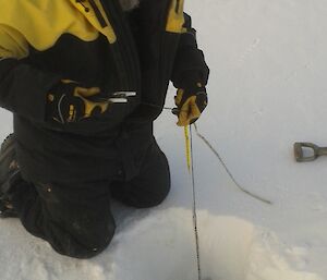 Rhys is holding a tape measure that is coming out of the hole he drilled in the sea ice. This allows him to measure the ice thickness.