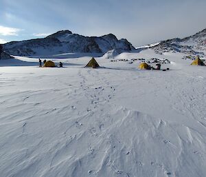 The winter camp site. On the horizon is the plateau where the wind barrels down towards Lake Druzhby. There are two polar pyramid tents and two polar domes. Four people are seen erecting the tents.