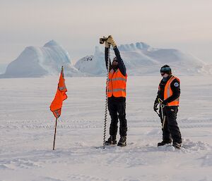 Sharky is holding a two metre long drill to take an ice core at a flagged post. Kirsten is holding the tape measure.