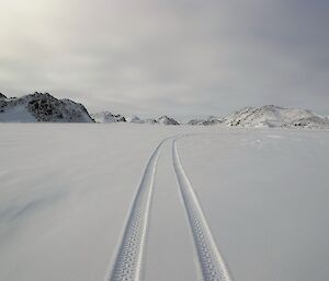 Quad tyre tracks are seen in the snow on the frozen fjord. The Vestfold Hills are in the background.