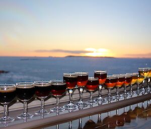 Sixteen small glasses containing beer ranging from black, to brown, to orange and gold in colour, sit on the Bar table with a view of sea ice and a sunset out the window.