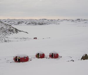 The red fibreglass melon and two apple huts are seen from a higher vantage point. Ellis fjord, frozen and covered in snow lies beyond the huts.