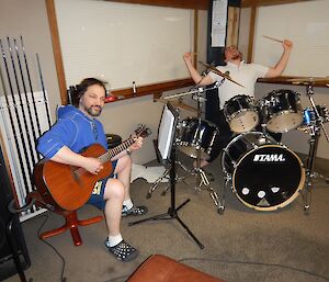 A man playing the drums and another playing the acoustic guitar.