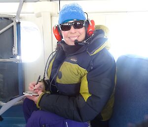 Smiling lady sitting on a plane wearing a beanie, headset and sunglasses holding a notebbok.