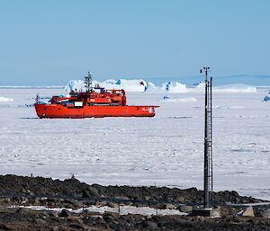 A red ship in sea ice with rocks and a communications pole in the foreground.
