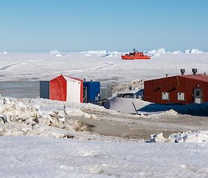 Buildings surrounded by snow with a red ship in the sea ice in the background.