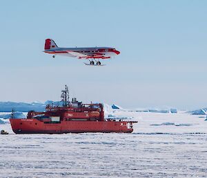 Red ship in sea ice with a small fixed wing plane flying above it.