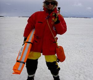 A man stands on the sea ice holding a fluro sea ice drilling kit.